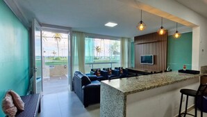 A modern kitchen with a central island, a dining area with a sofa, and a TV mounted on the wall. - COSTE0114 - Inba Stella Residence (Salvador)