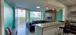 A modern kitchen with a central island, a dining area with a sofa, and a TV mounted on the wall. - COSTE0114 - Inba Stella Residence (Salvador)