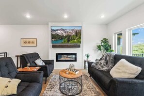 A living room with a fireplace, a large TV mounted above it, and a view of mountains through the window. - Luxurious Lakefront Townhouse Near Deer Valley Resort (Heber City)