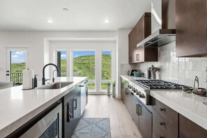 A modern kitchen with a large island, stainless steel appliances, and a view of a green landscape through large windows. - Luxurious Lakefront Townhouse Near Deer Valley Resort (Heber City)