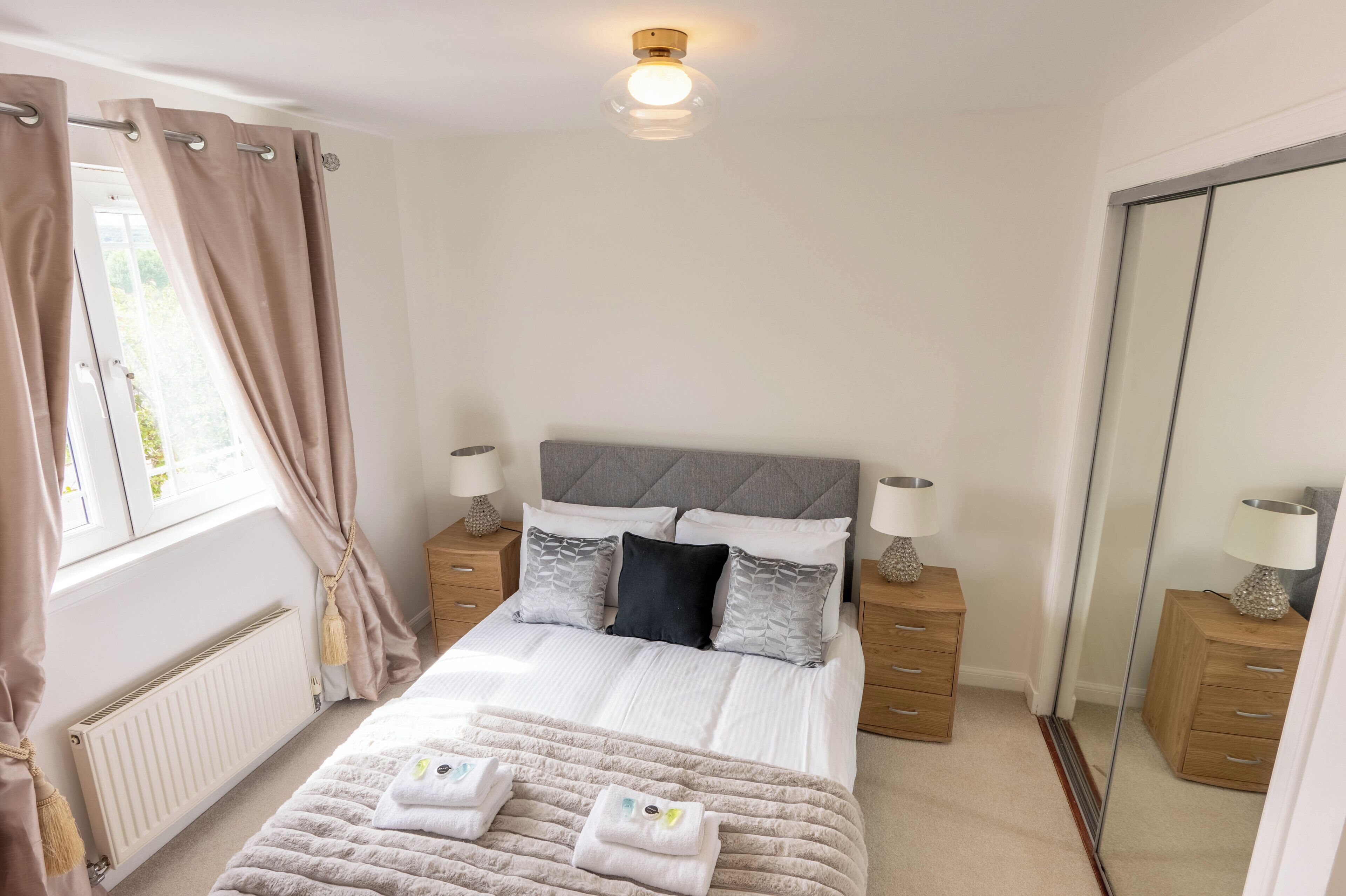 Quarto com cama, mesas de cabeceira, espelho, radiador e janela com cortinas.