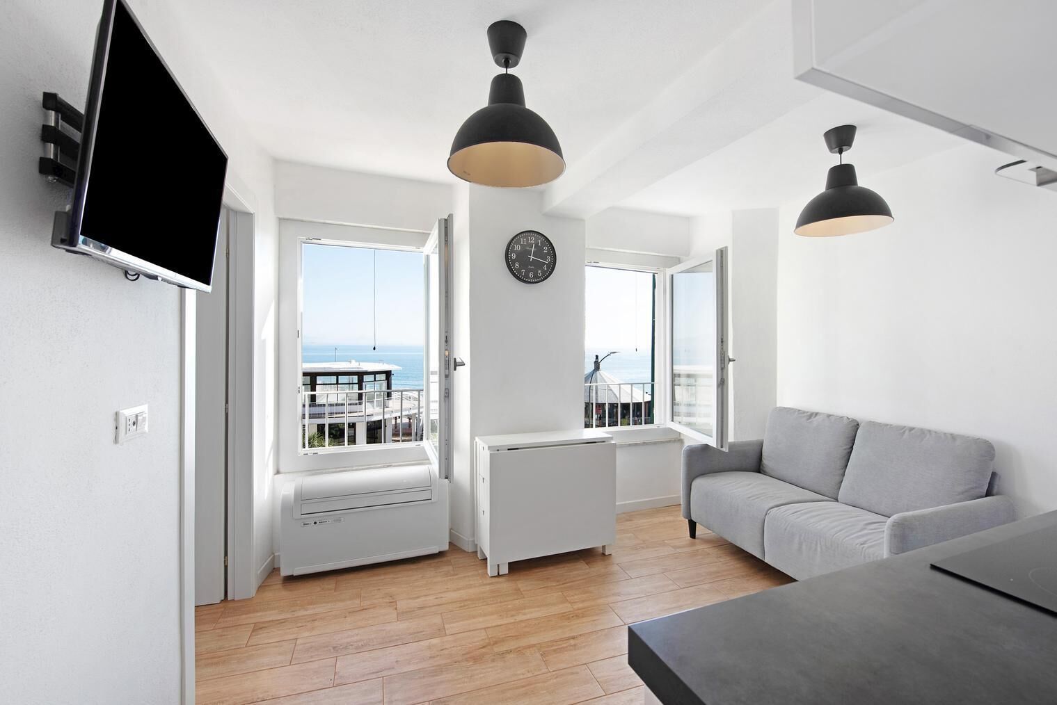 Una sala de estar moderna con un sofá gris, un televisor de pantalla plana montado en la pared, un amplio ventanal con vistas al mar e iluminación colgante.