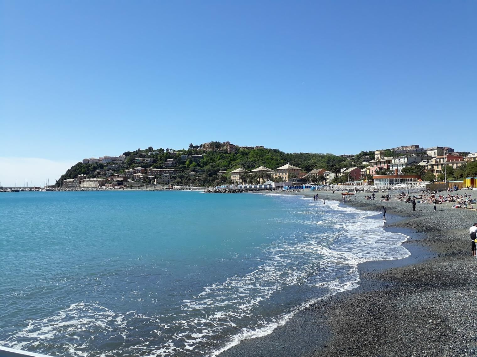 A coastal beach with a rocky shoreline, buildings in the background, and clear blue water.