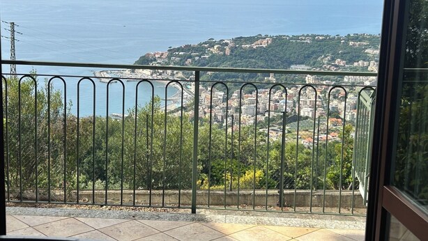 A balcony with a metal railing overlooking a coastal cityscape with buildings and greenery.