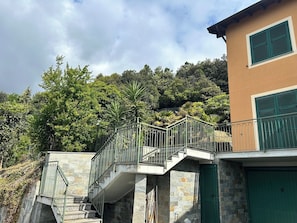 Eine Steintreppe führt zu einem Balkon mit Metallgeländern, einer Steinsäule und einem Gebäude mit grünen Fensterläden.