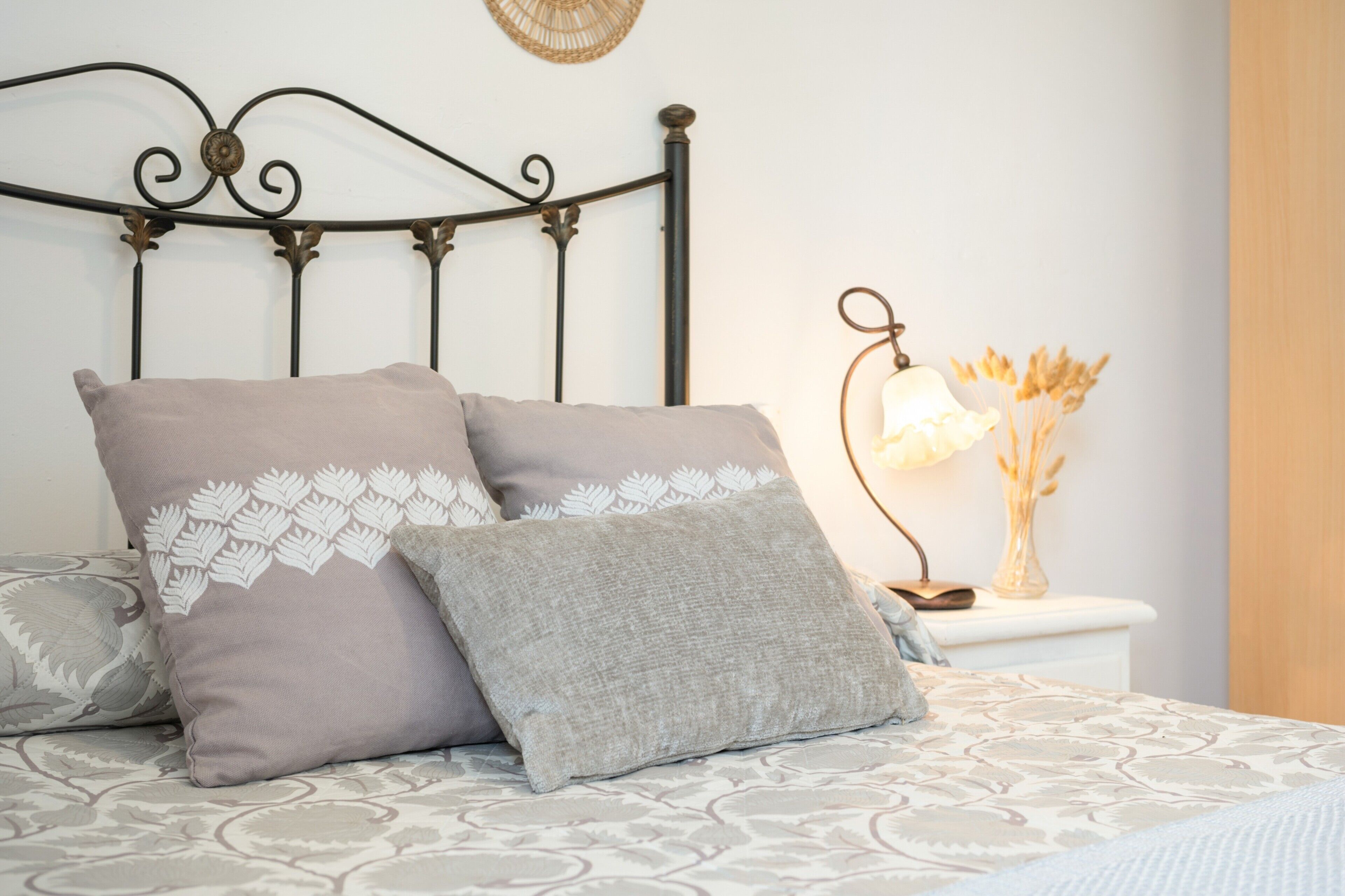 Un lit avec une couvre-lit à motifs, deux oreillers ornés de motifs de feuilles, une tête de lit en métal, une table de chevet avec une lampe et un vase contenant des fleurs séchées.