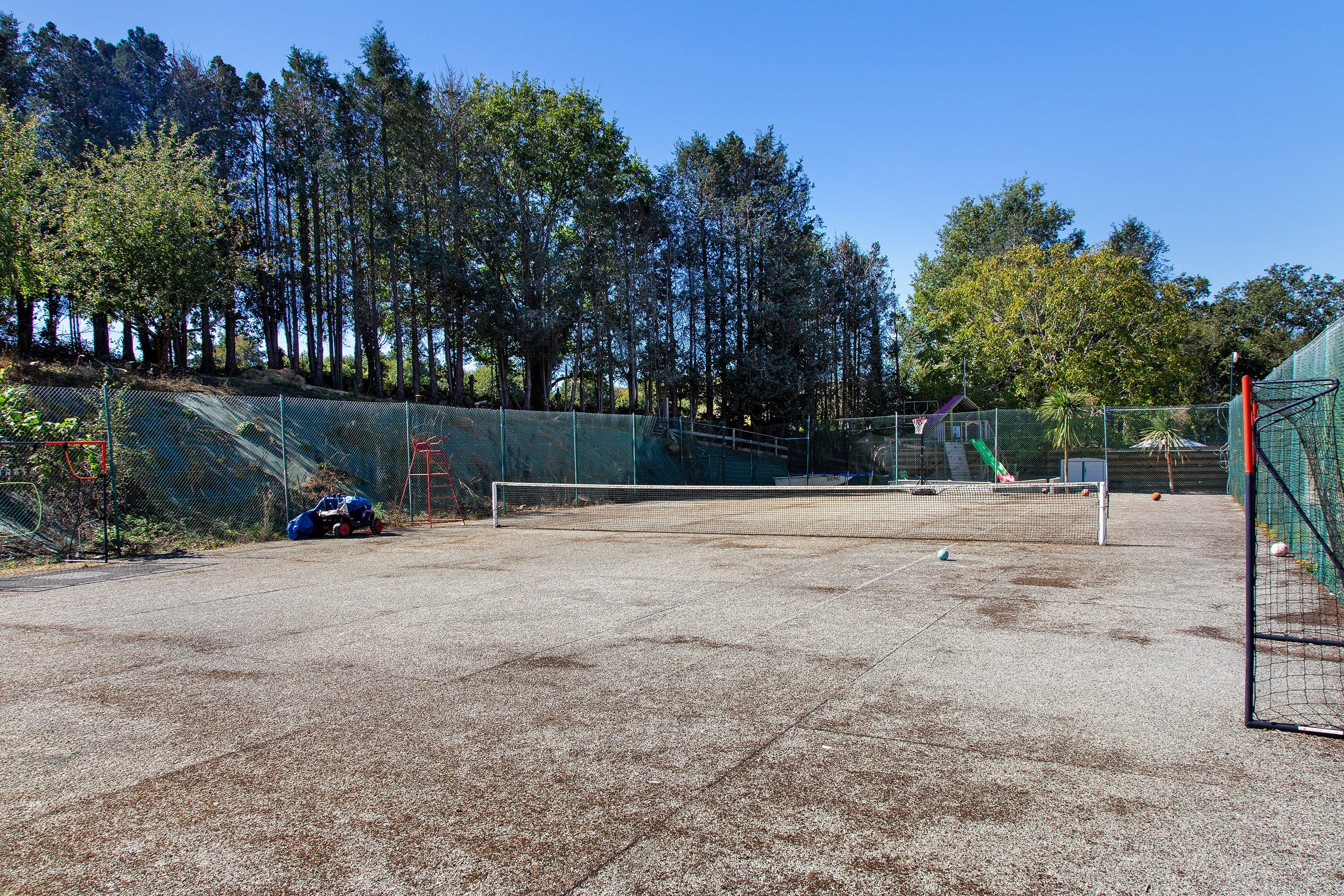 An outdoor tennis court with a net, surrounded by a fence and trees.