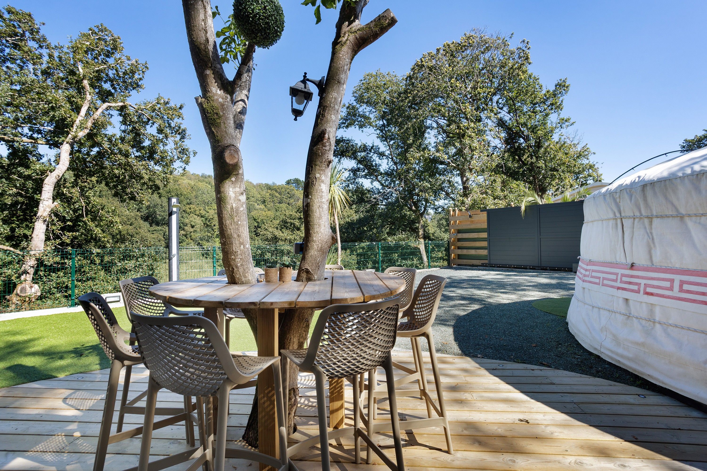 An outdoor dining area with a round wooden table and wicker chairs, surrounded by trees and a fenced garden.