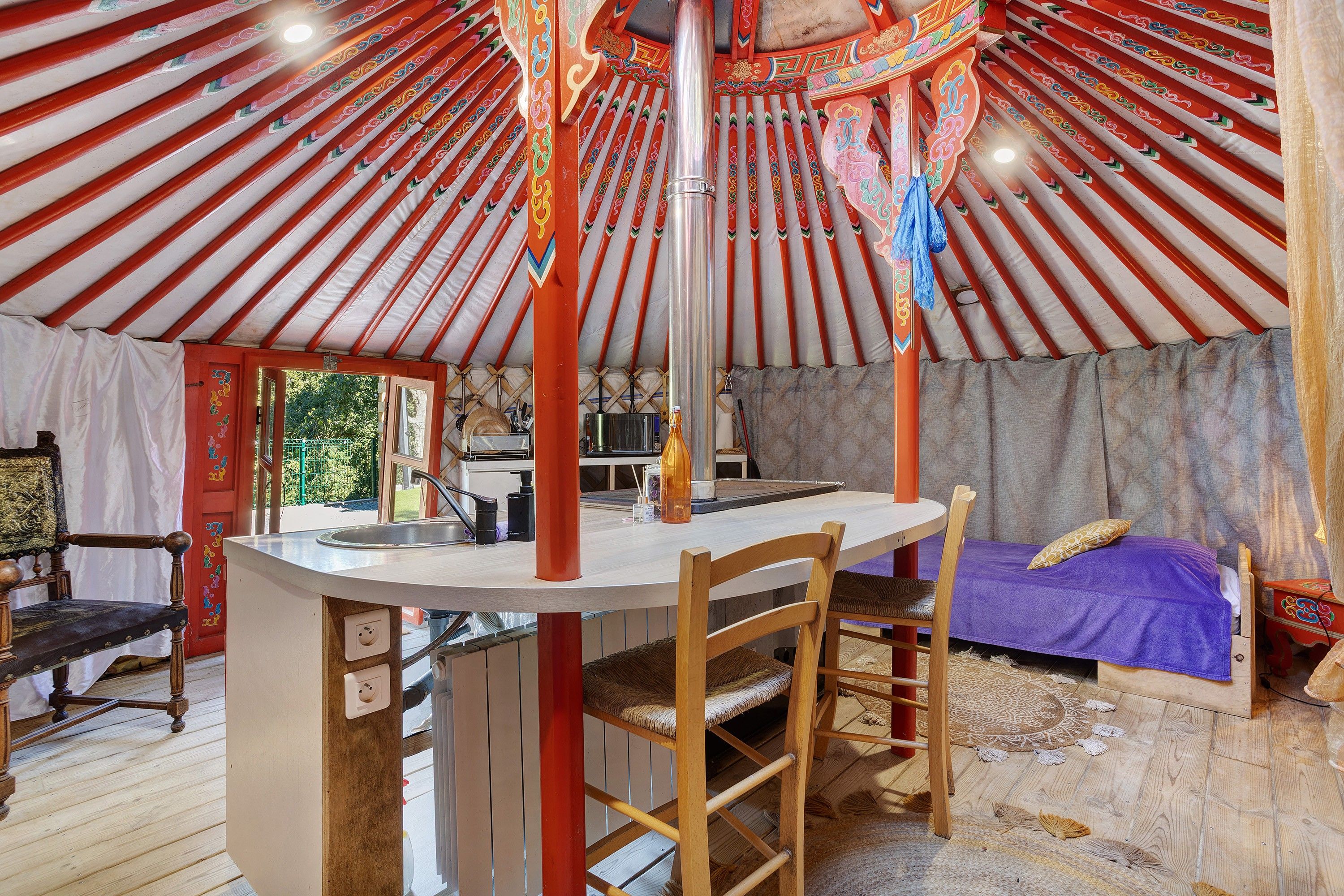 A yurt interior with a central wooden table, a bed with a purple bedspread, and a chair.