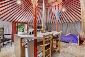 L’intérieur d’une yourte, avec une table en bois au centre, un lit recouvert d’une couverture violette et une chaise.