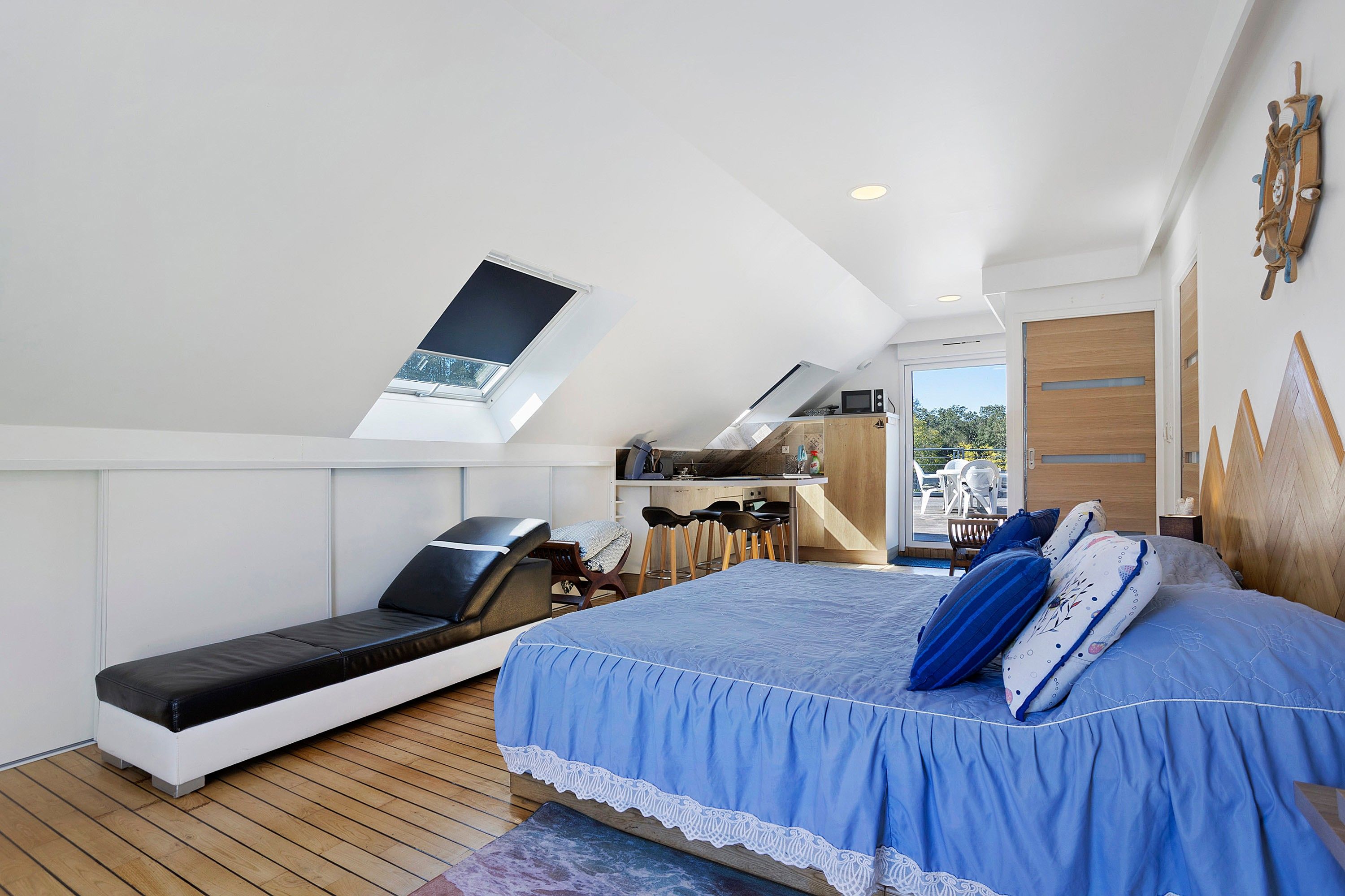 A bedroom with a skylight, a bed with blue bedding, a small seating area, and a kitchenette.