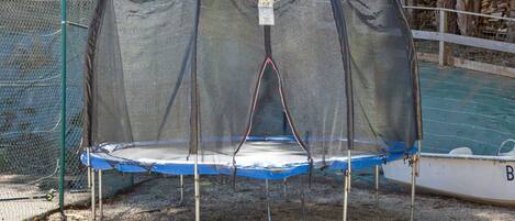 Un trampoline doté d’un filet de protection, entouré d’une clôture en treillis métallique, et un bateau portant la lettre « B » à l’arrière-plan.