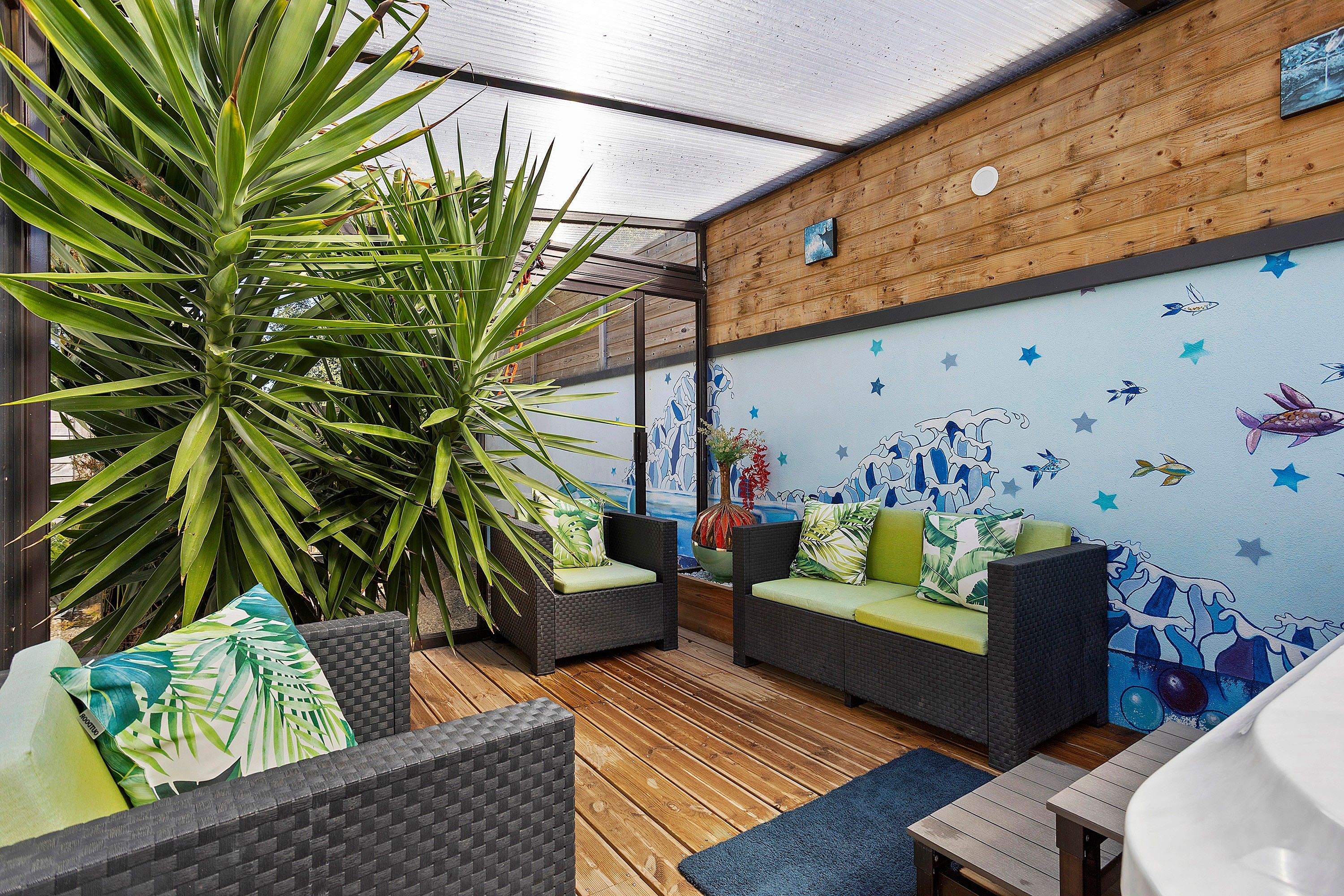 Un espacio al aire libre con mesas y sillas de mimbre, una planta grande y un mural en la pared.