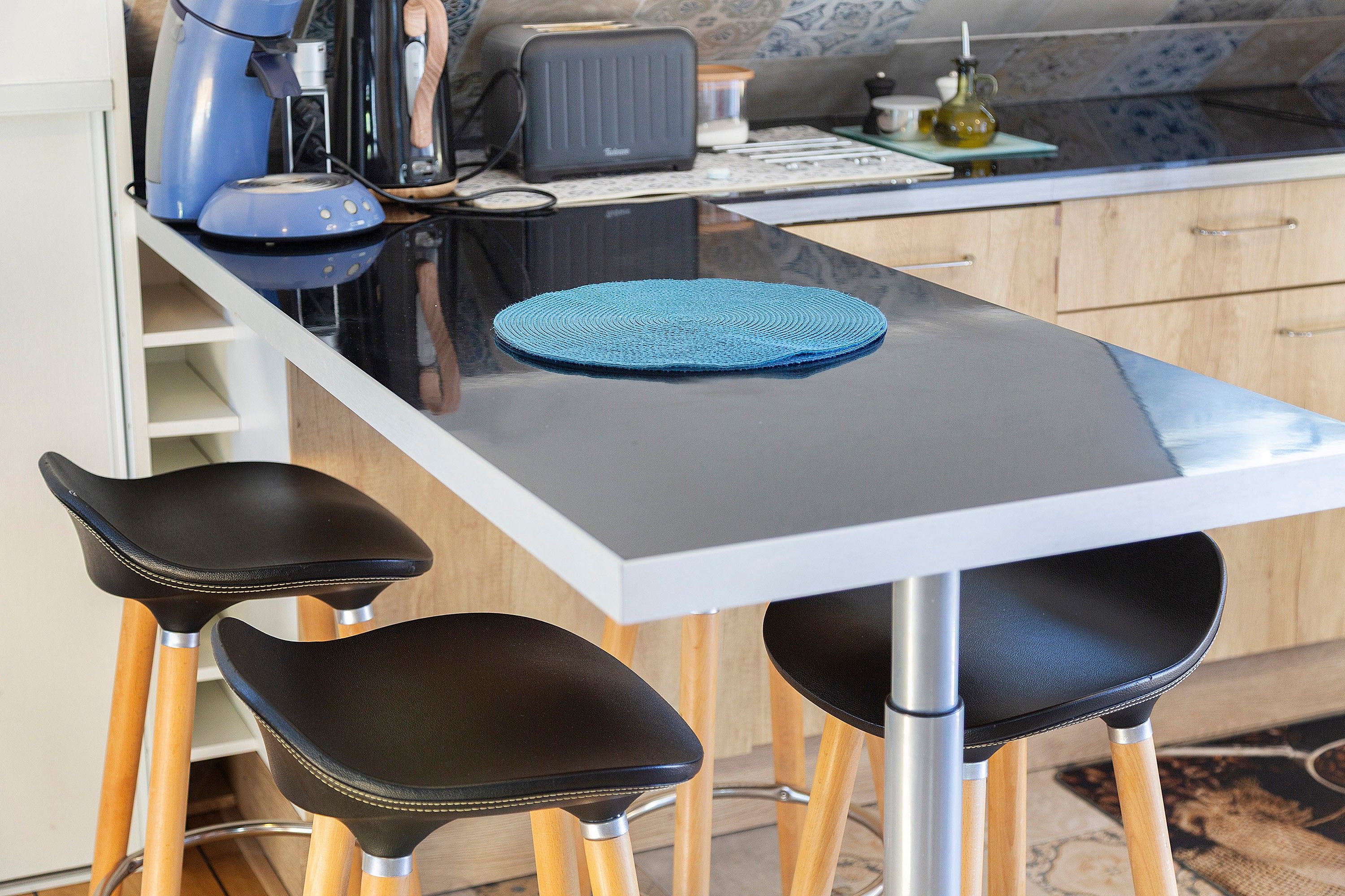 A modern kitchen with a white countertop, two black bar stools, a blue woven placemat, and a coffee machine.