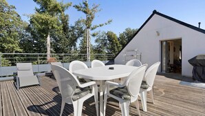 An outdoor patio with a round table and white chairs, surrounded by a wooden deck and a white building with a sloped roof. - In Nature – Spa – 2km from City Center (Quimper)