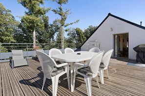 Een terras buiten met een ronde tafel en witte stoelen, omgeven door een houten terras en een gebouw met een schuine witte daklijn.