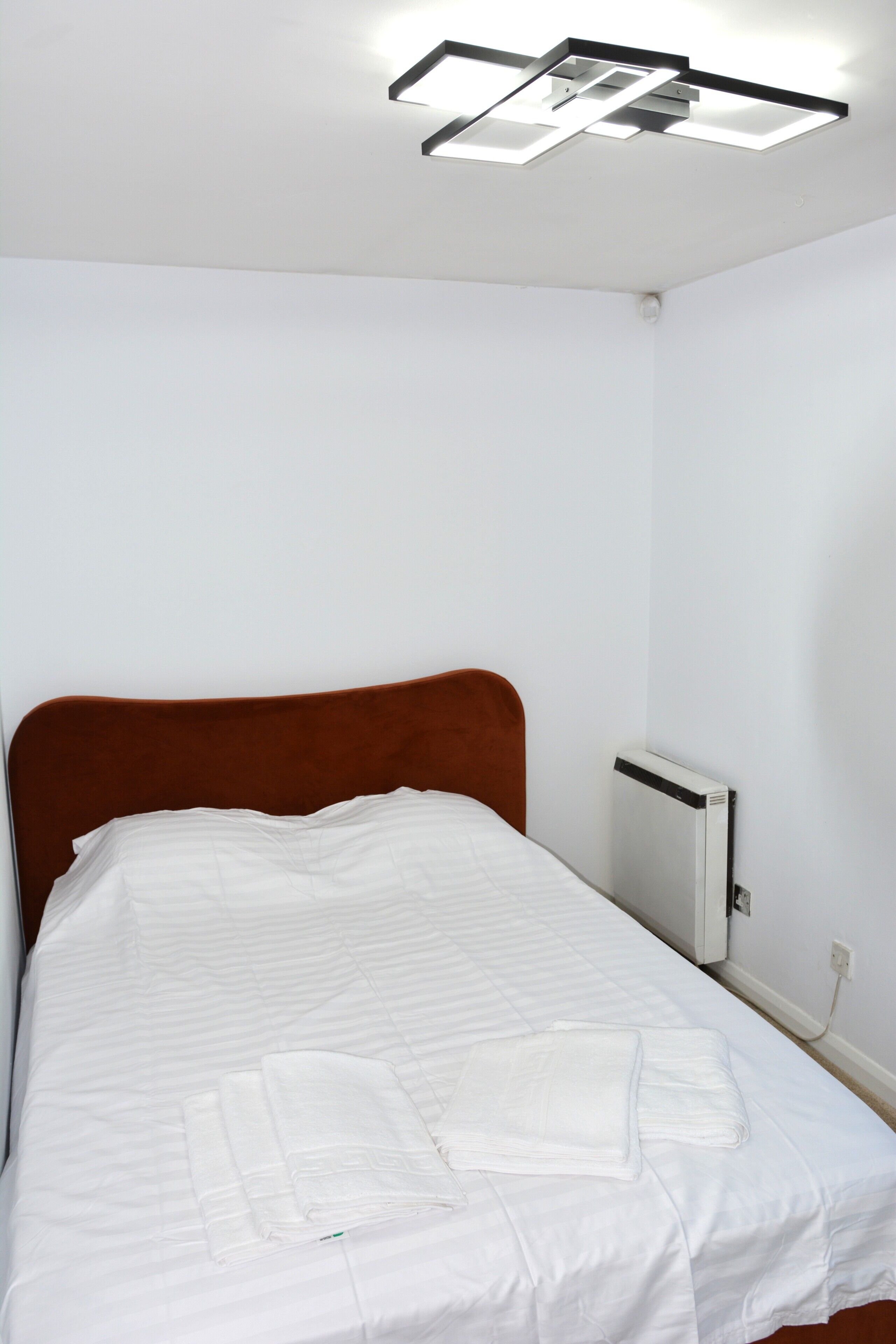 Una cama individual con cabecera marrón, ropa de cama blanca y un radiador blanco en una esquina.