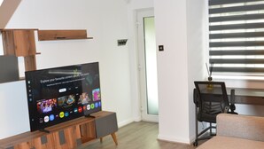 A modern living room with a flat-screen TV, a wooden coffee table, a grey sofa, and a window with blinds. - Magic House- London (London)