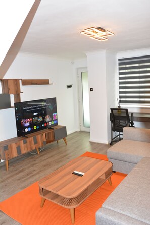 A modern living room with a flat-screen TV, a wooden coffee table, a grey sofa, and a window with blinds. - Magic House- London (London)