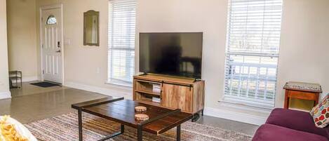 A living room with a sofa, a coffee table, a television on a wooden stand, and two windows with blinds.