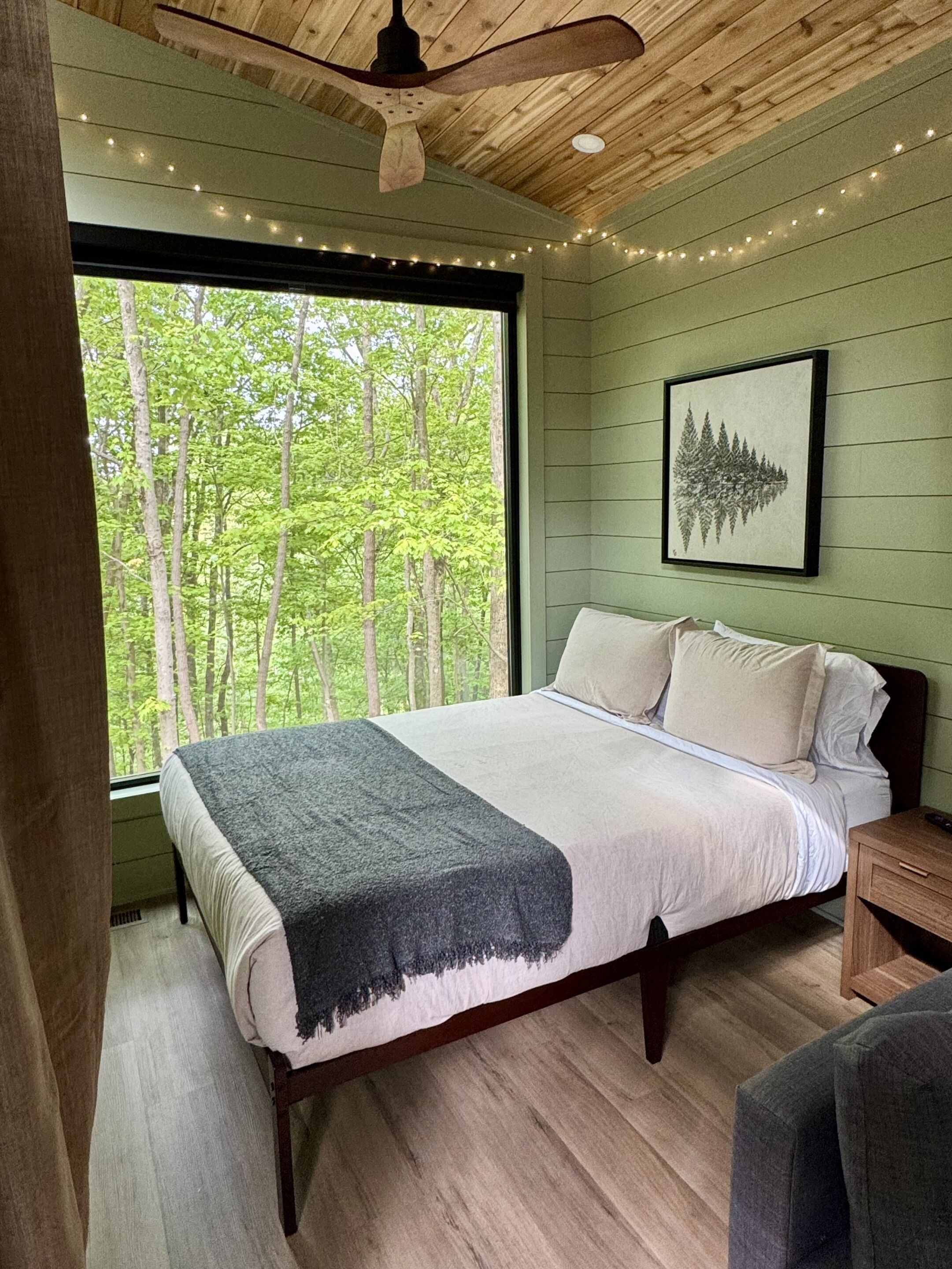Une chambre avec une grande fenêtre, un lit recouvert d’une couverture, un ventilateur de plafond en bois et un tableau encadré accroché au mur.