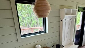 Et rum med et træbord og stole, en hængende lampe, et vindue med udsigt til træer, og et hvidt opbevaringsskab.