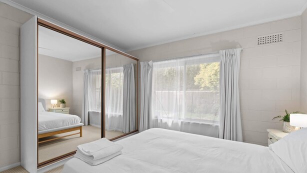 A bedroom with a large mirror, a bed with white linens, a bedside table with a lamp, and a window with sheer curtains. - Courtyard 2 Bed Apartment Mile End (Mile End)