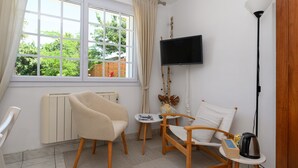 Une pièce avec un téléviseur, deux chaises, une petite table, un radiateur et une fenêtre avec des rideaux.