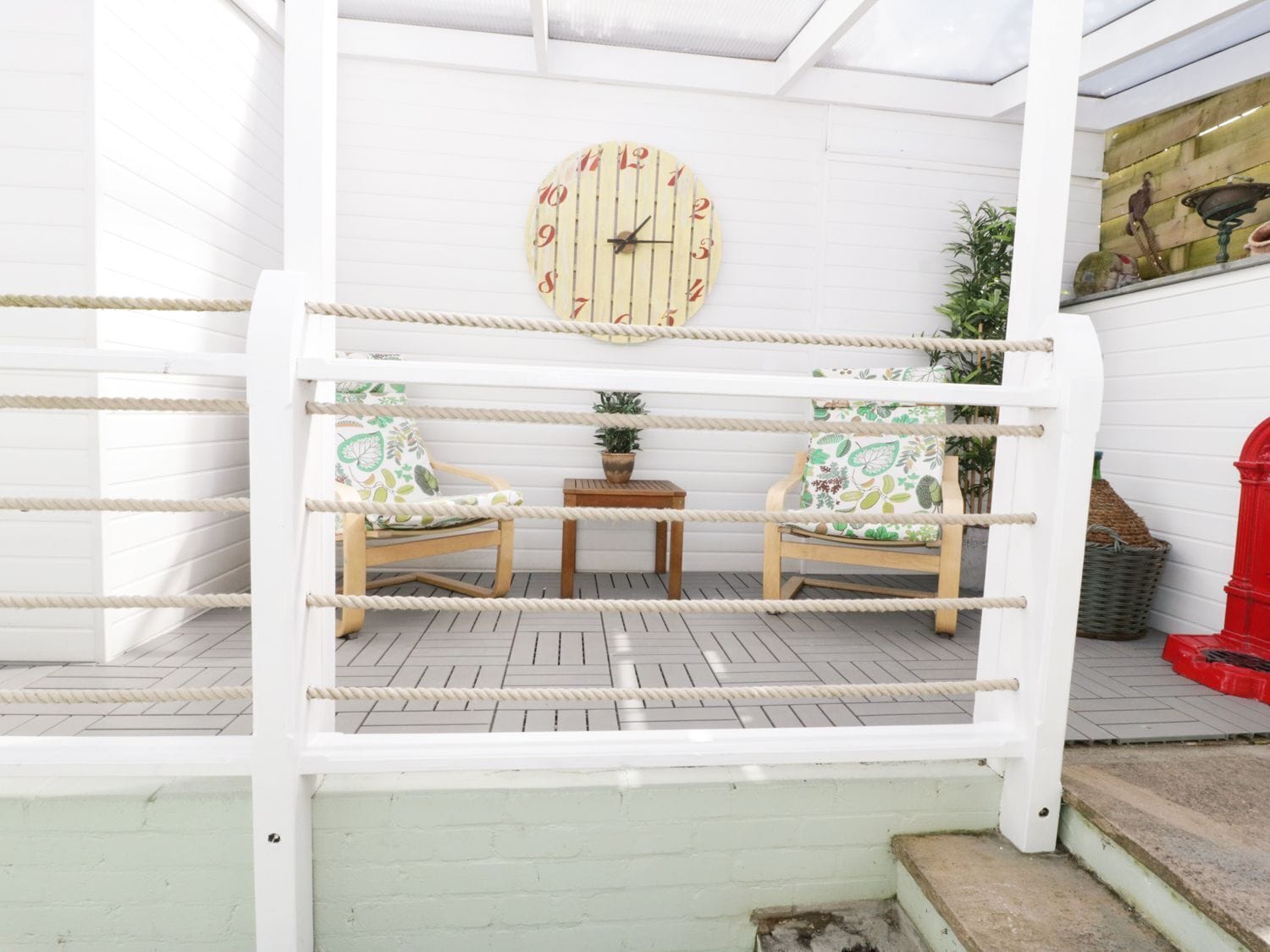 Un patio abrité comprenant deux chaises en bois, une petite table et une horloge fixée au mur.