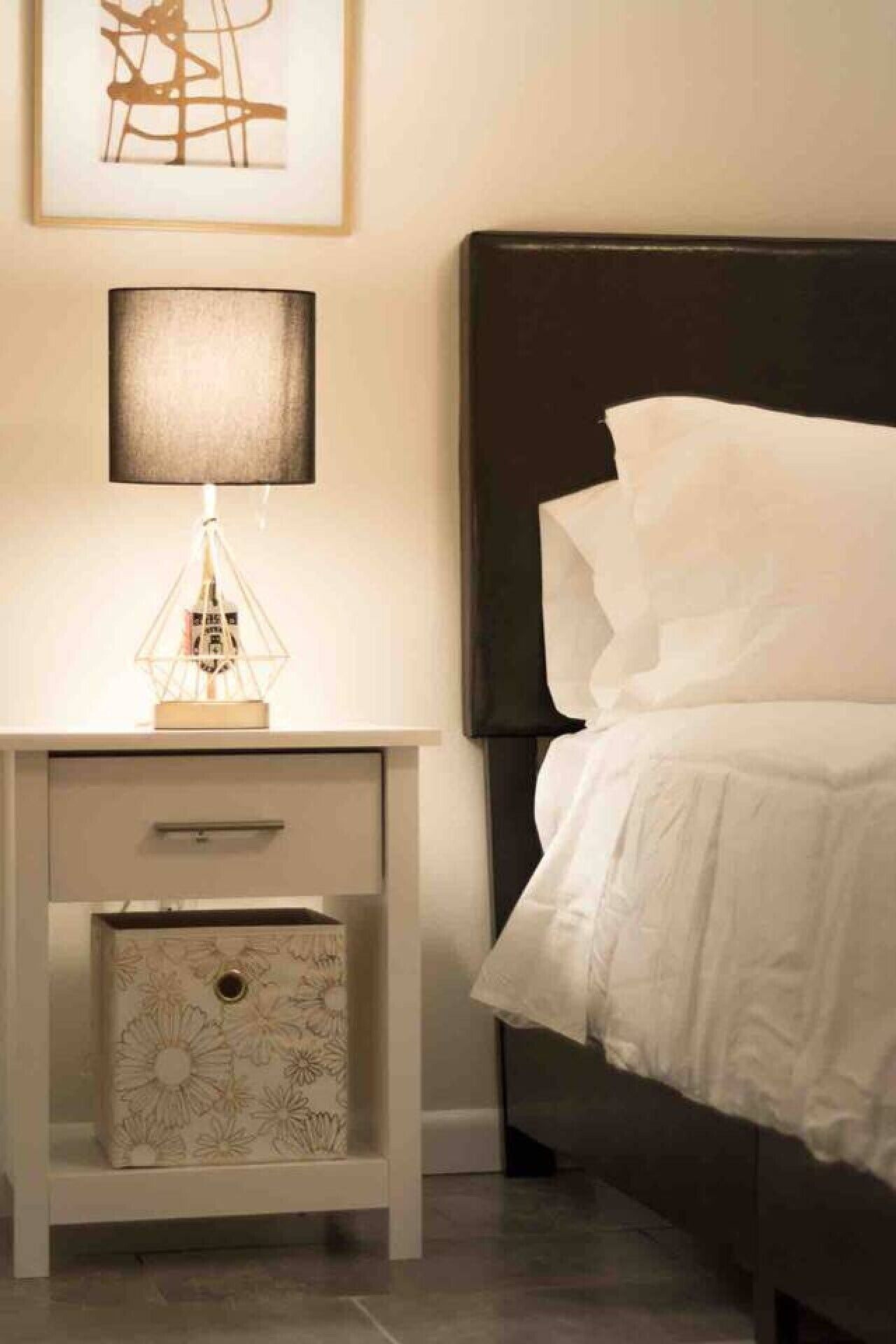 A neatly made bed with white bedding, a bedside table with a lamp, a framed abstract artwork, and a patterned storage box.