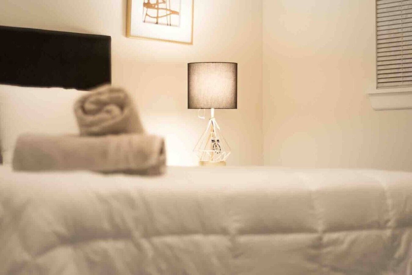 Una cama bien hecha con una toalla doblada, una lámpara de noche y un cuadro en la pared.