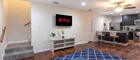 A modern living room with a flat-screen TV, a staircase, a kitchen area, and a blue patterned rug.