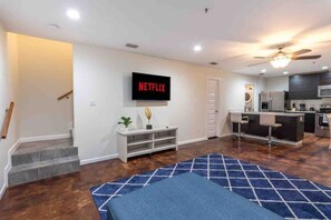 A modern living room with a flat-screen TV, a staircase, a kitchen area, and a blue patterned rug.