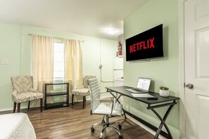 Ein Zimmer mit einem Bett, zwei gemusterten Sesseln, einem Schreibtisch mit einem Laptop, einem Fernseher, der 'NETFLIX' anzeigt, und einem Feuerlöscher.