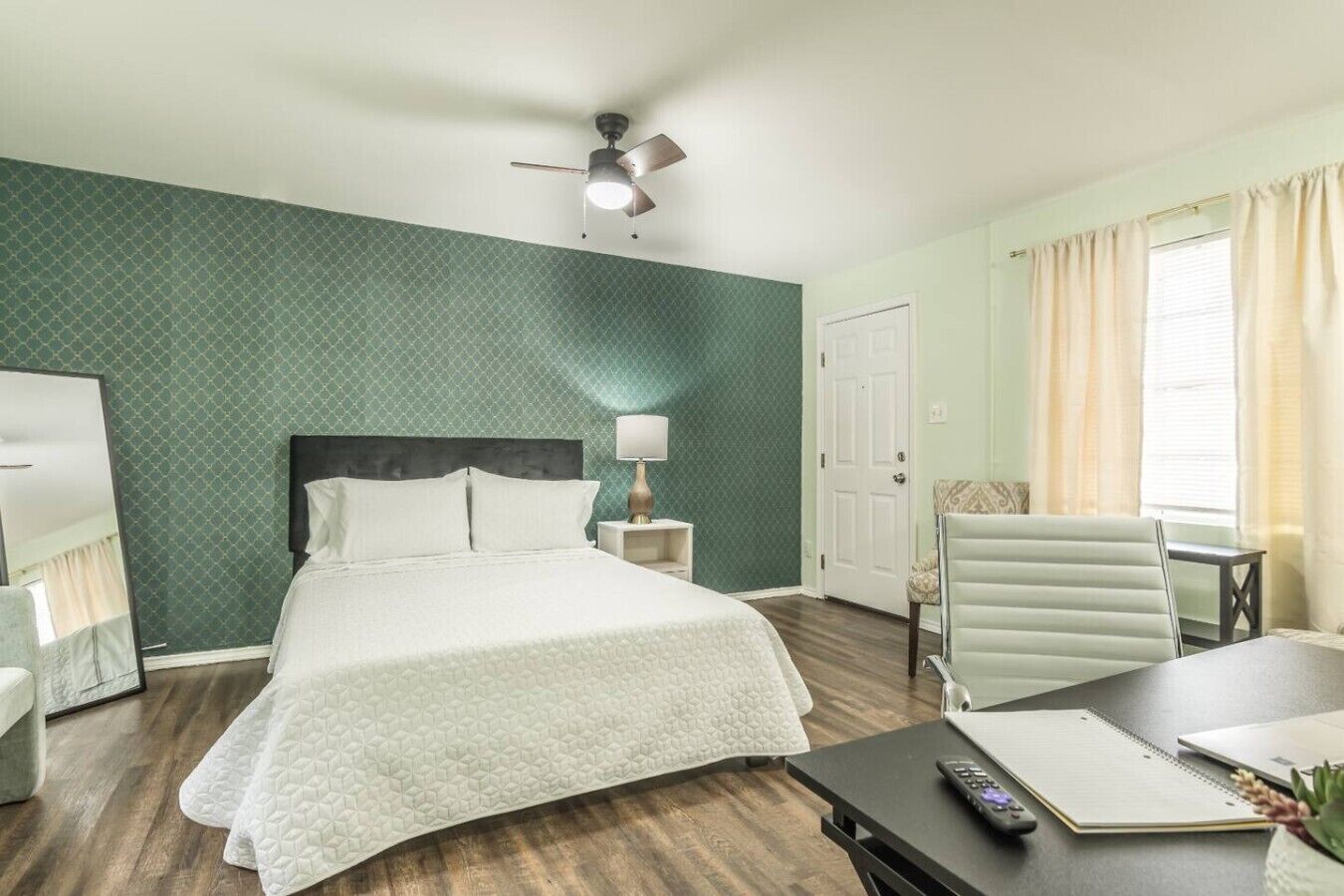 A hotel room with a bed, a desk with a chair, a lamp, a ceiling fan, and a window with curtains.