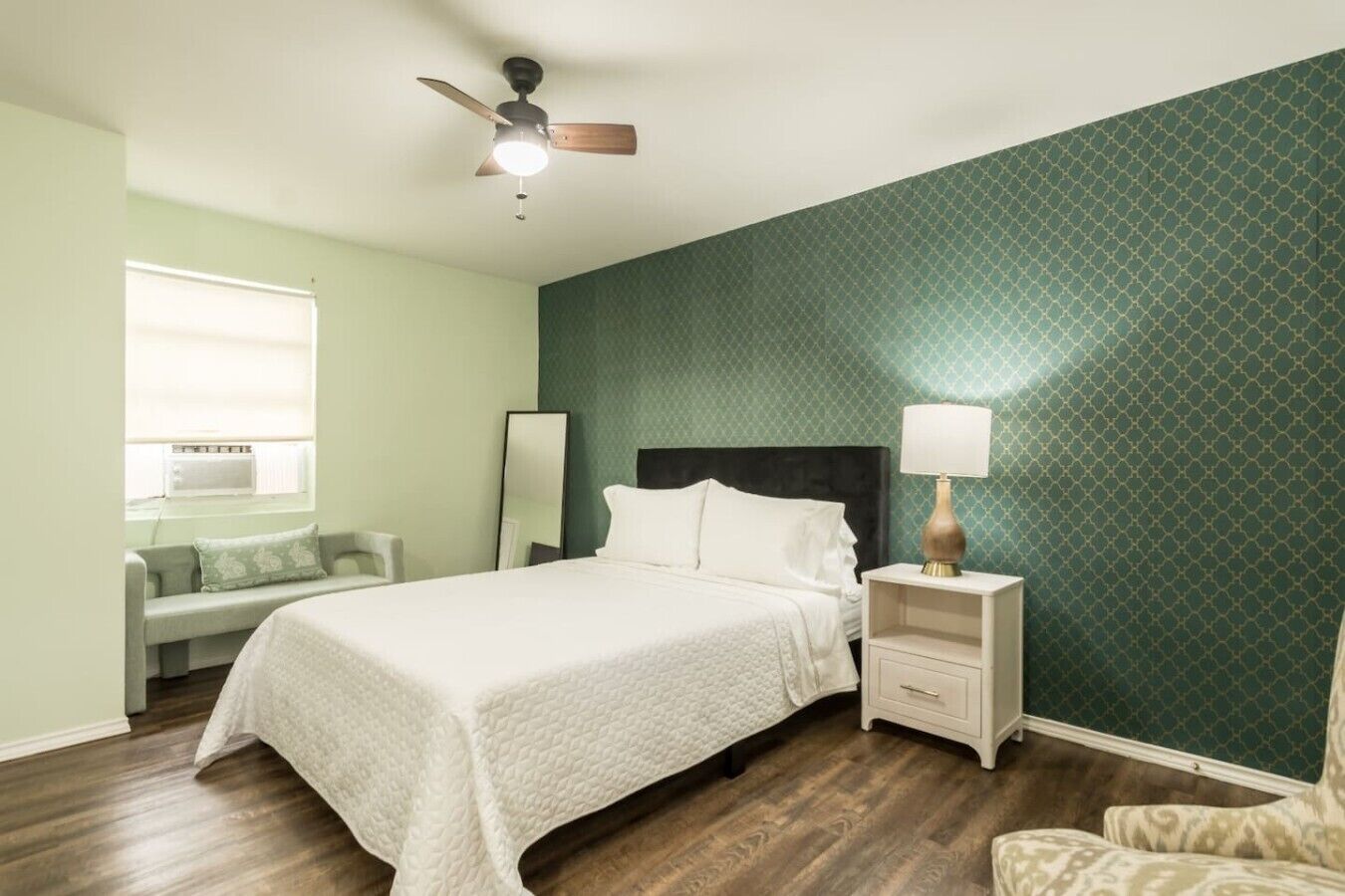 Un dormitorio con cama, mesitas de noche, lámpara, ventilador de techo, ventana y pared con diseño geométrico.