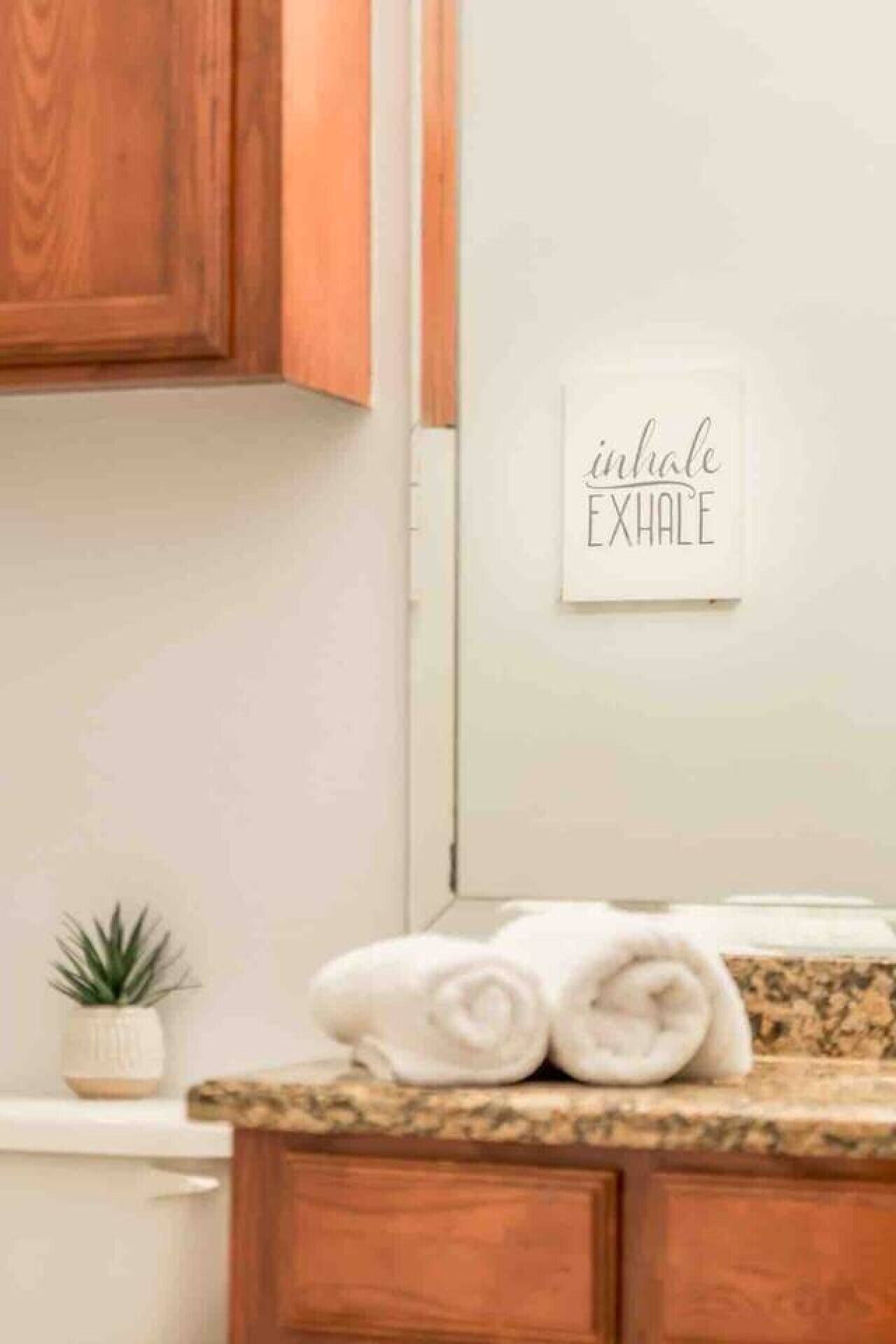 A bathroom with wooden cabinets, a towel rack with folded towels, a small potted plant, and a wall-mounted sign with the words 'inhale EXHALE'.