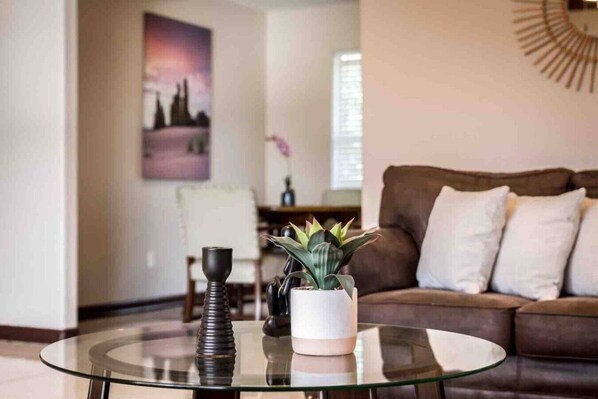 A living room with a glass coffee table, a brown sofa, a potted plant, and a painting on the wall.