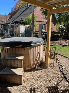 A wooden hot tub with steps, a wooden pergola, and a chair in a backyard with gravel and a brick building in the background.