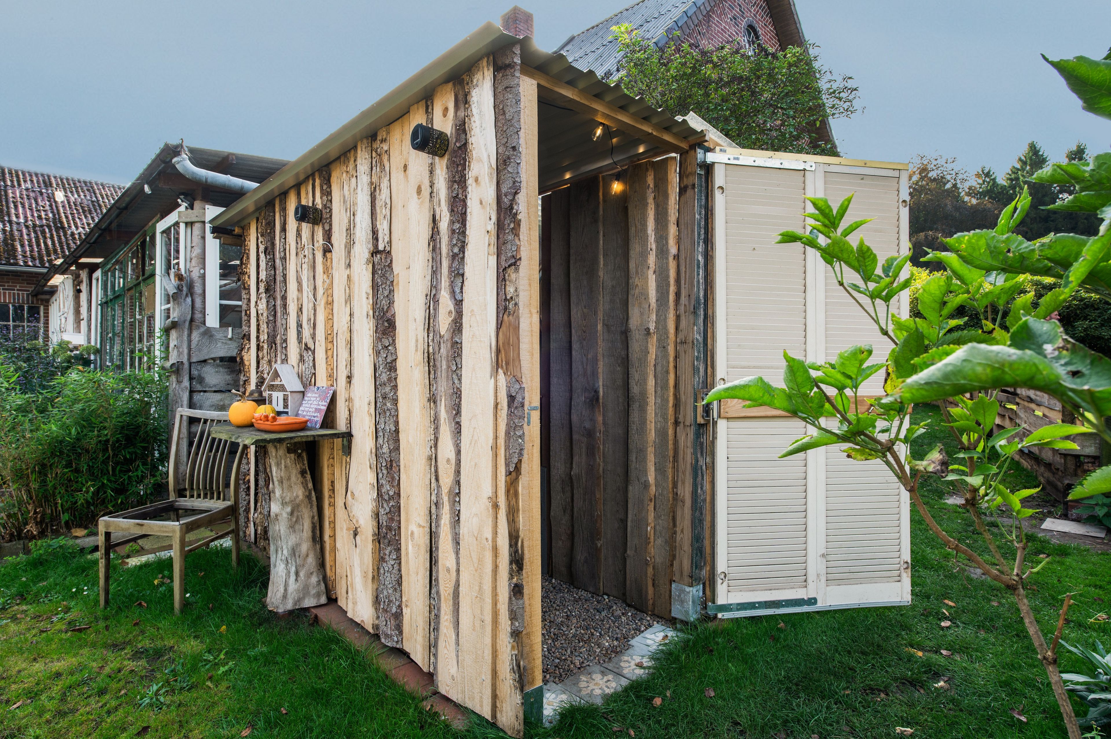 Ein hölzernes Gartenhaus mit Tür, ein kleiner Tisch mit Kürbissen und ein Stuhl davor.