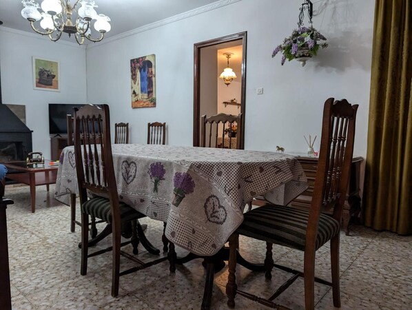 Ein Esszimmer mit einem für vier Personen gedecktem Tisch, umgeben von Holzsesseln. Über dem Tisch hängt eine Chandelier, an der Wand hängt ein Gemälde und eine Tür führt in einen anderen Raum.