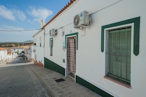 A narrow street with a white building featuring green trim, a window with metal bars, and an air conditioning unit. - Casa Rural Villagarcia – Comfort and Privacy in El Real de la Jara (El Real de la Jara)