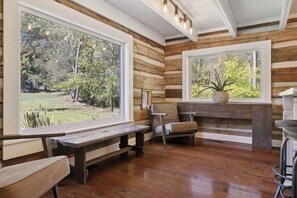 Interior rústico de una cabaña con paredes de madera, ventanas grandes, un banco y una silla.