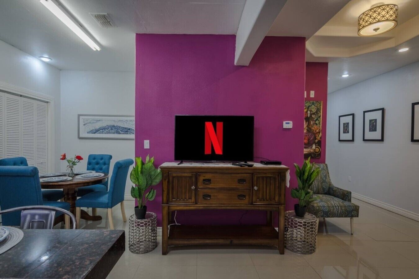 Uma sala de estar com televisão fixada numa parede roxa, um armário de madeira, cadeiras azuis e uma mesa de jantar.