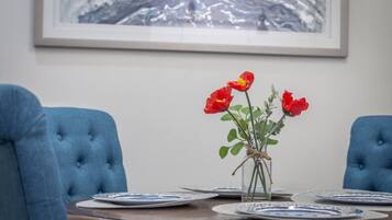 Una taula de dinar amb cadires blaves tapissades, un ram de flors vermelles en un vas i un quadre al mur.