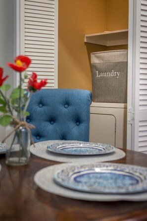 Et spisebord med tallerkener, en blå polstret stol og en vasketøjskurv mærket 'Laundry' i et rum med hvide persienner.