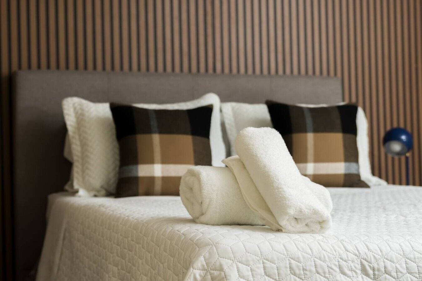 A neatly made bed with a quilted cover, two rolled towels, and two pillows with a plaid pattern.