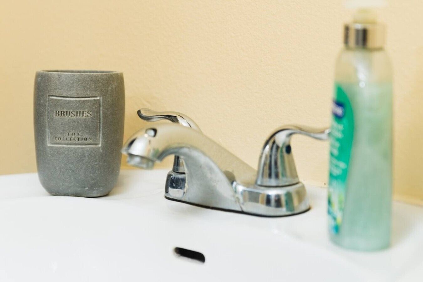 A bathroom sink with a faucet, a soap dispenser, and a container labeled 'BRUSHES THE COLLECTION'.