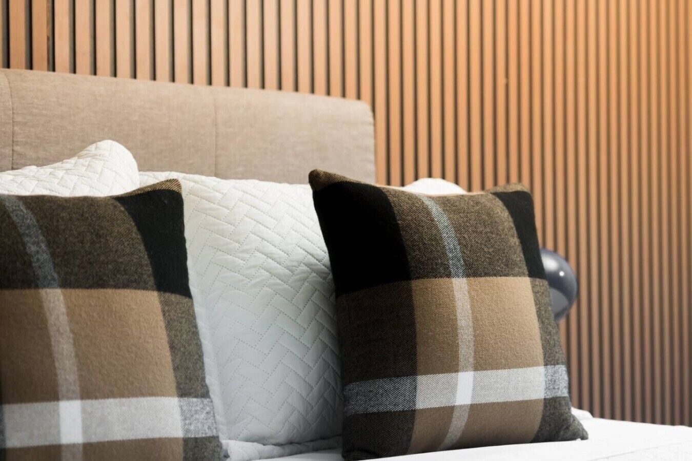 Una cama con manta y almohadas a cuadros, cabecera de madera y pared con listones verticales.