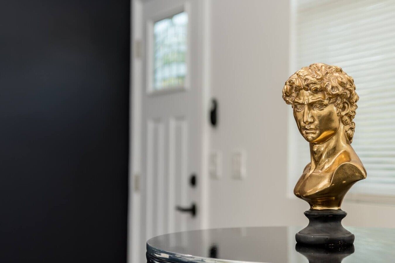 A bust sculpture on a reflective surface in a room with a door and blinds in the background.
